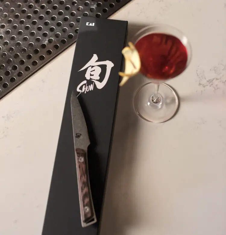 A Kanso paring knife lies on a Shun knife box next to an amber-hued cocktail.